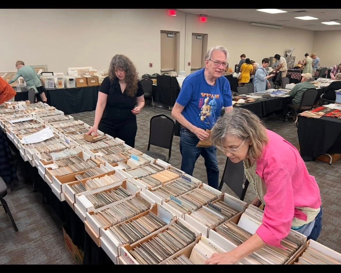 Three collectors looking through long rows of postcard boxes on a dealer table.
