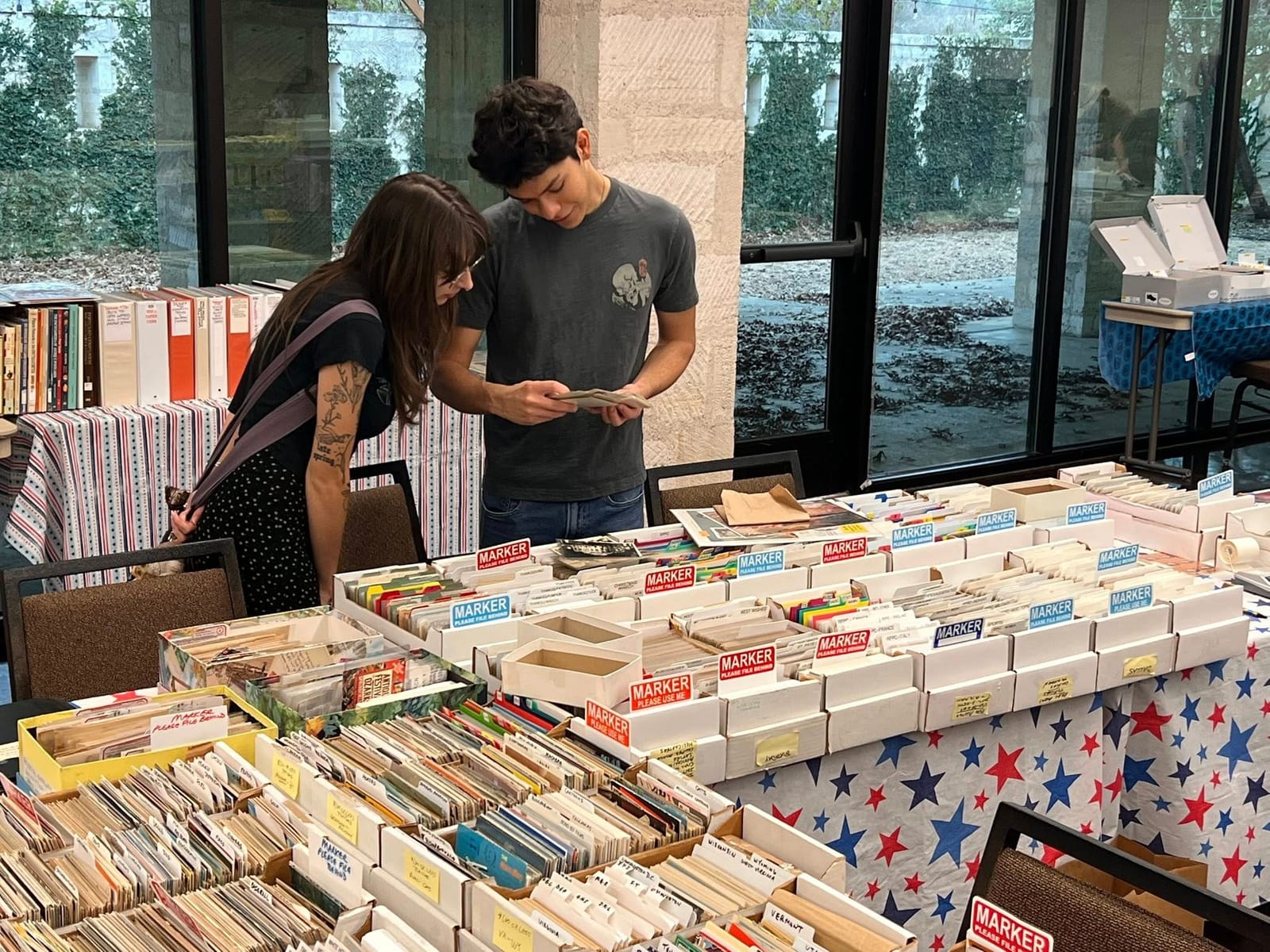 A couple examining a postcard together at a table of boxes by the windows.
