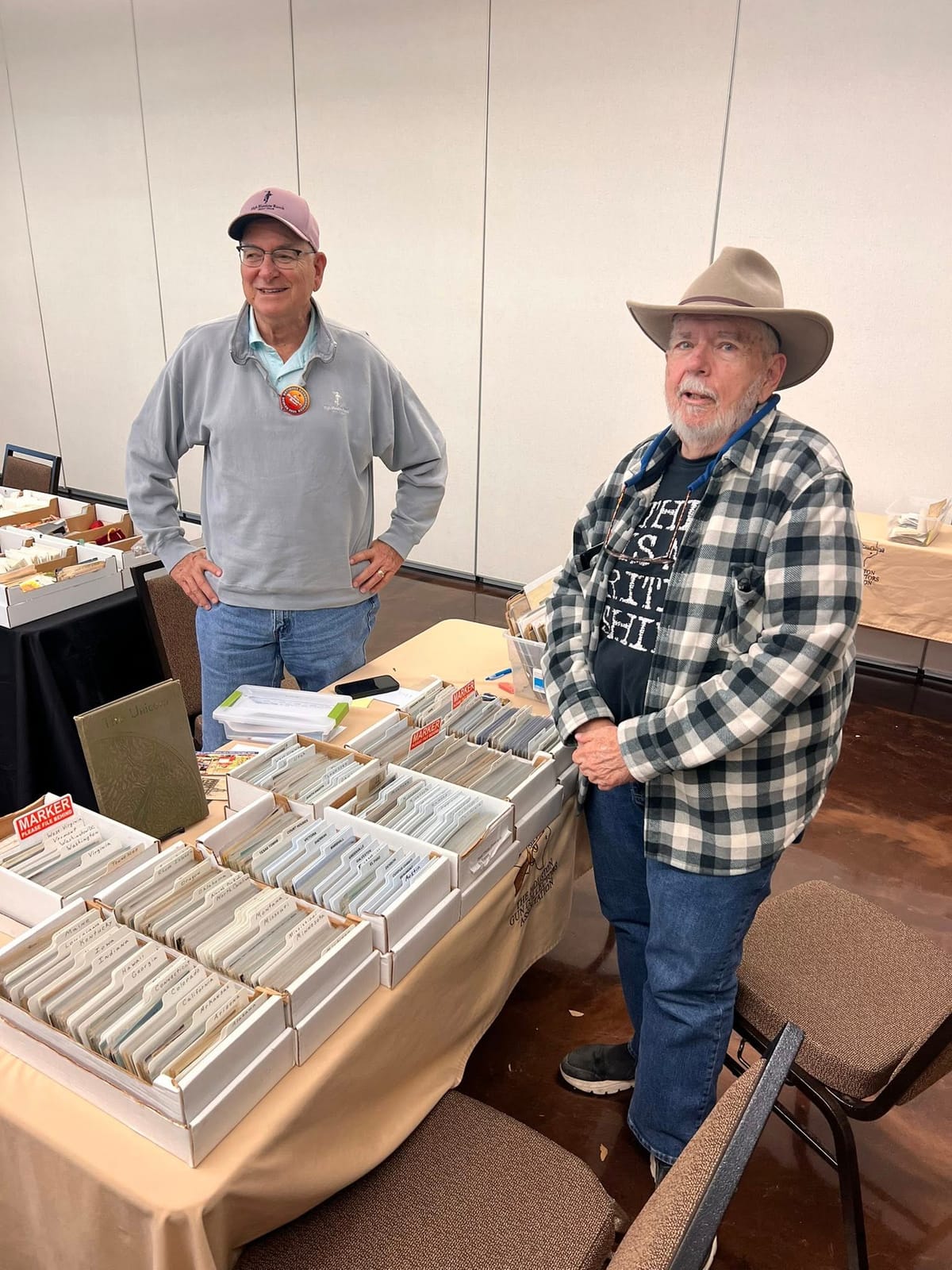 Two collectors at a table of postcards organized state by state, one wearing a cowboy hat.
