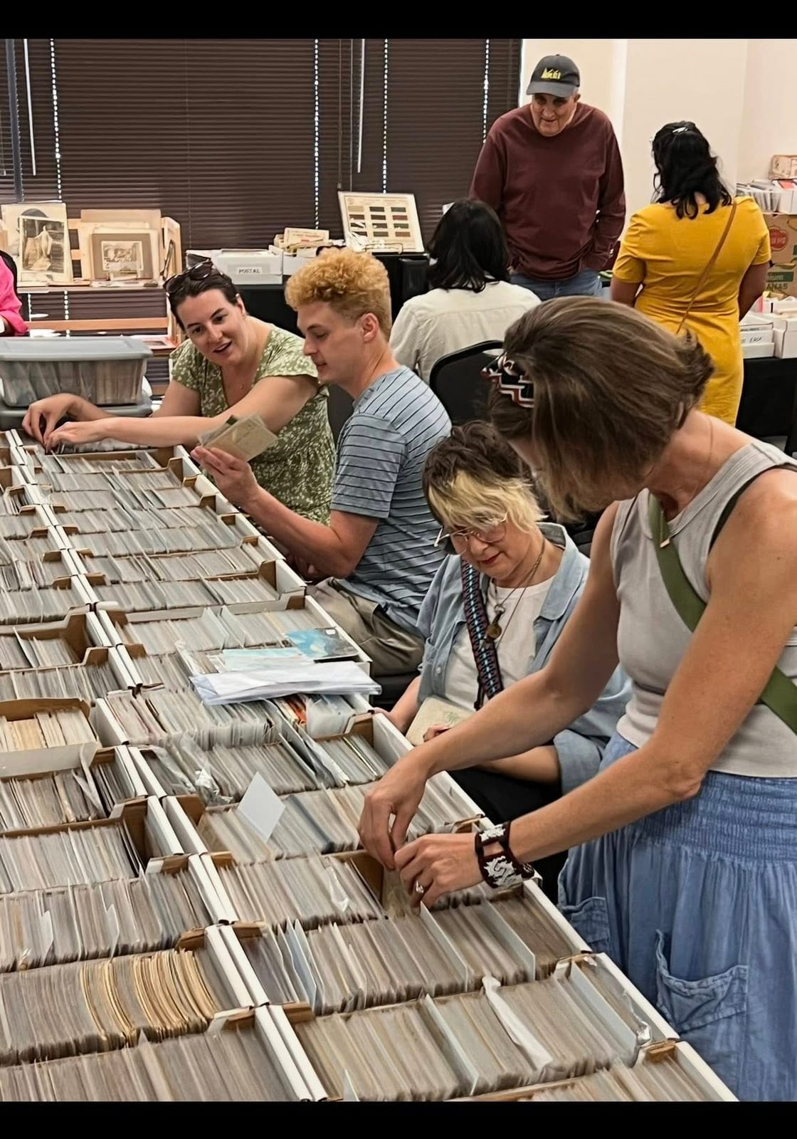 Several people closely browsing long rows of postcard boxes at the show.