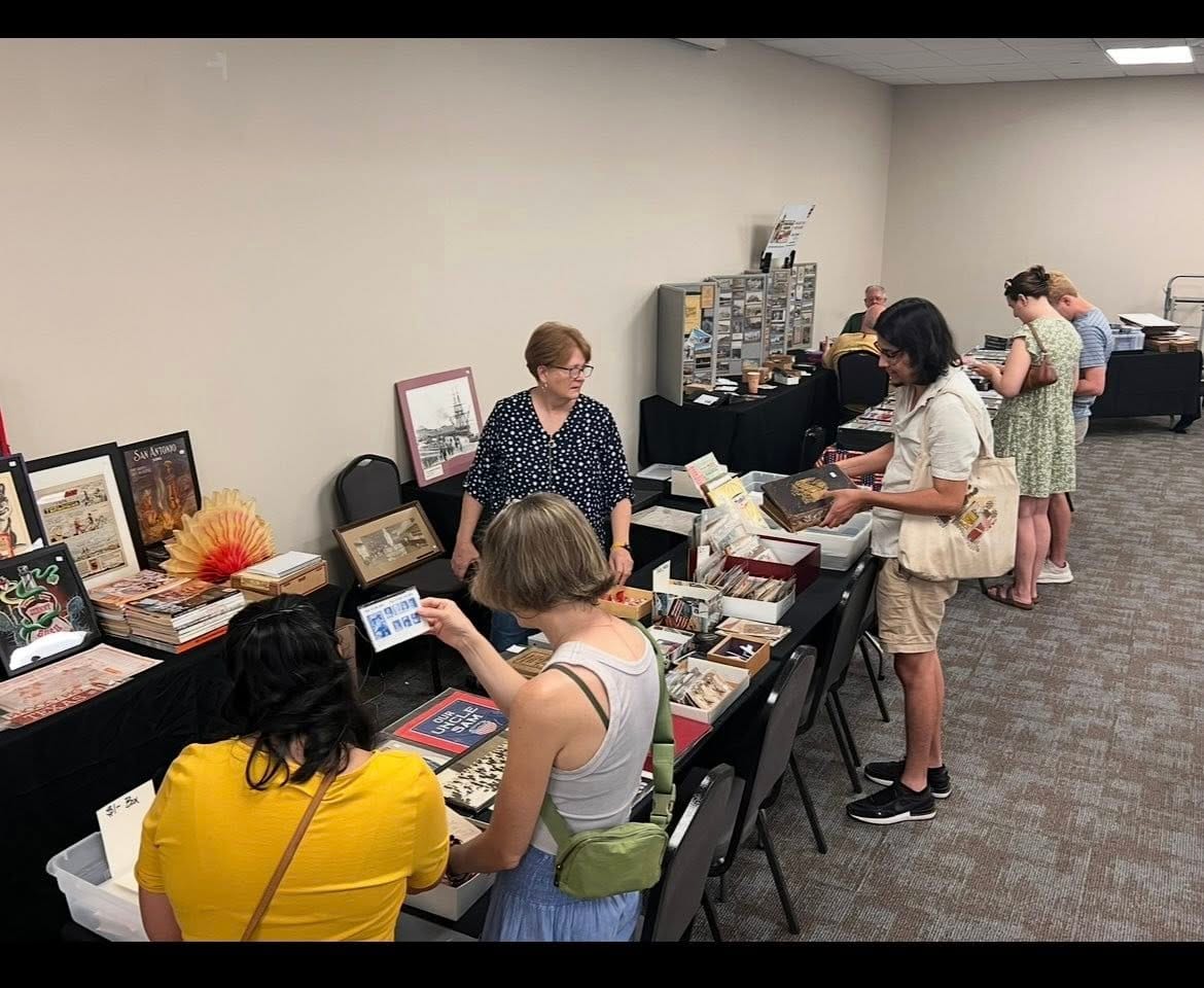 A dealer table with framed prints and boxes of paper ephemera as attendees browse.